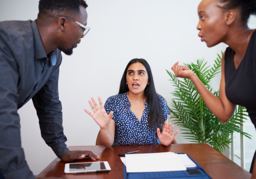 A woman sitting at a desk trying to break up an argument between a man and woman who are standing either side of the desk.